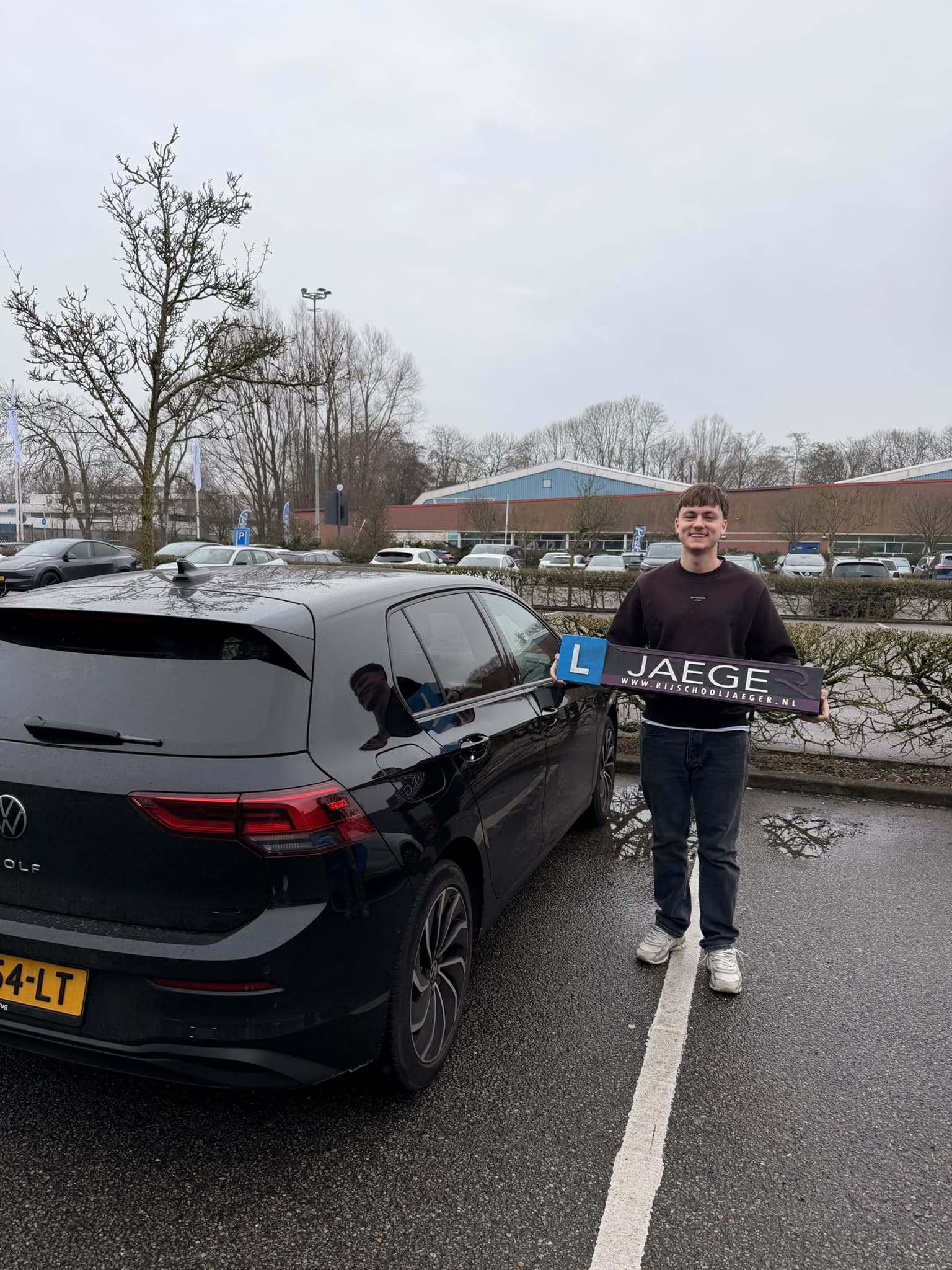 Geslaagde leerling met een Jaeger-bord bij de zwarte Volkswagen-lesauto